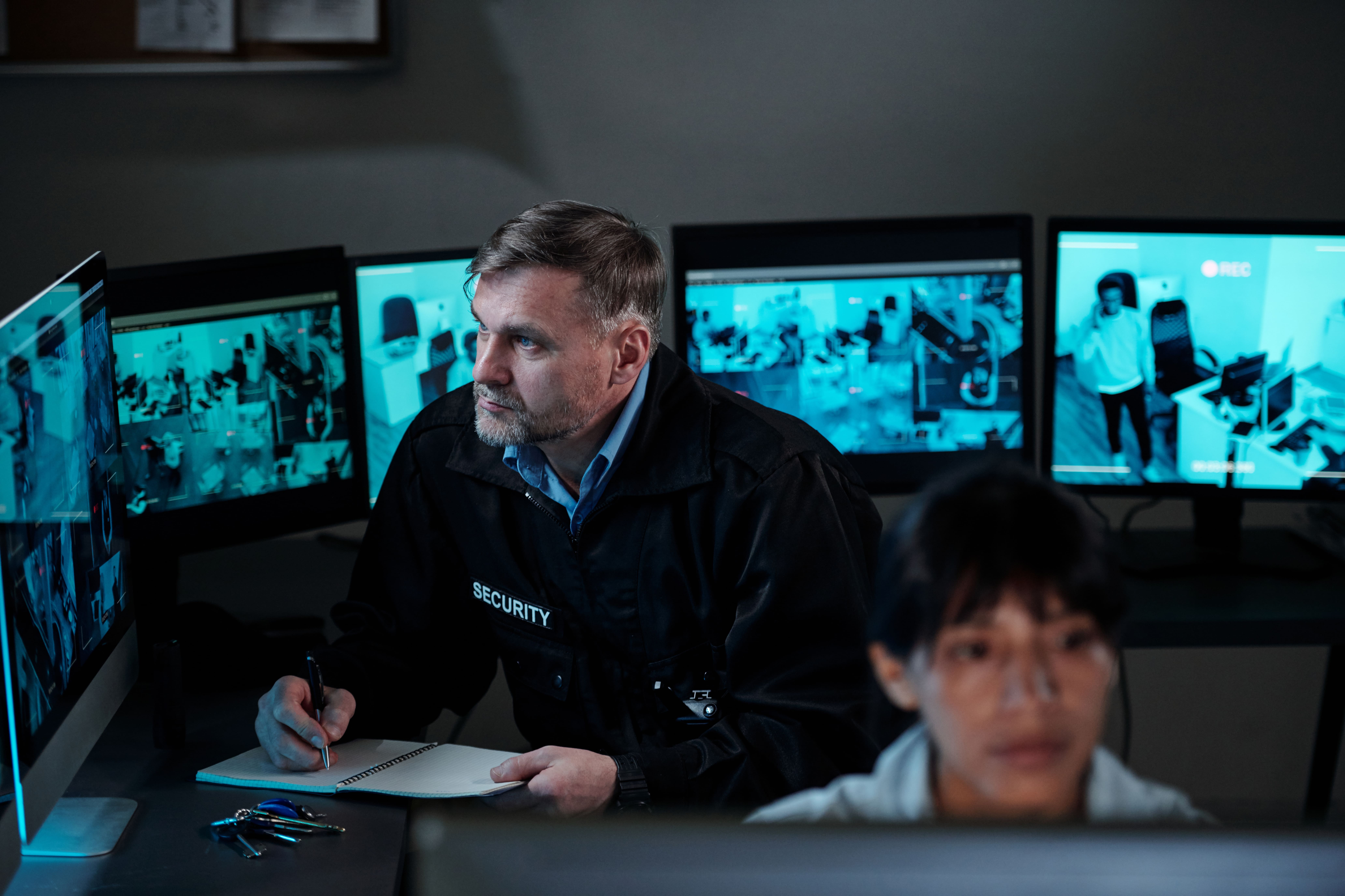Two security personnel are monitoring multiple surveillance screens in a dimly lit control room.