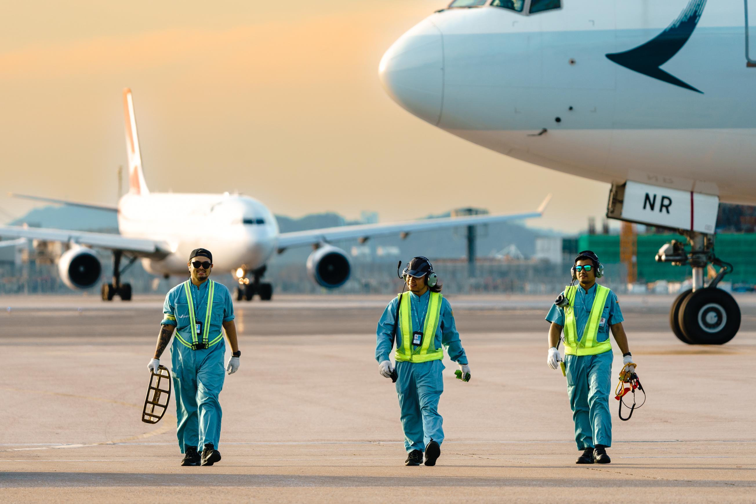 Three happy Aircraft Engineering Licence Trainees at HAECO walking and smiling with airplane and sunset in the background